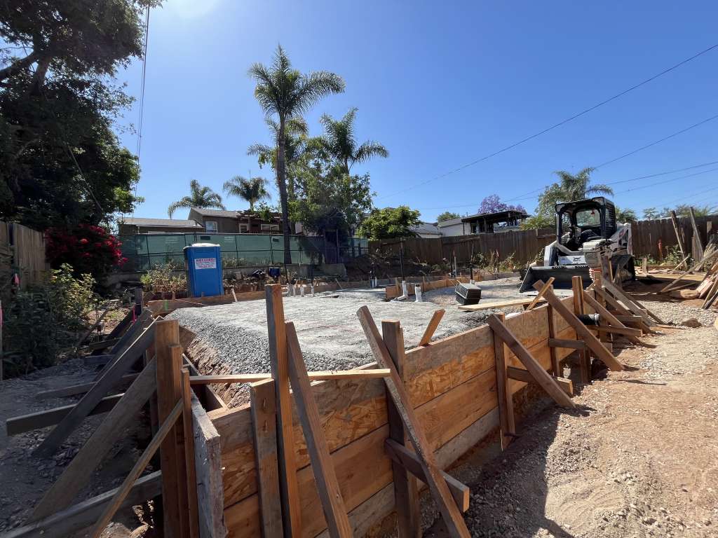 Wider view of the prepared construction site including gravel-filled foundation, wooden forms, portable toilet, and light equipment.