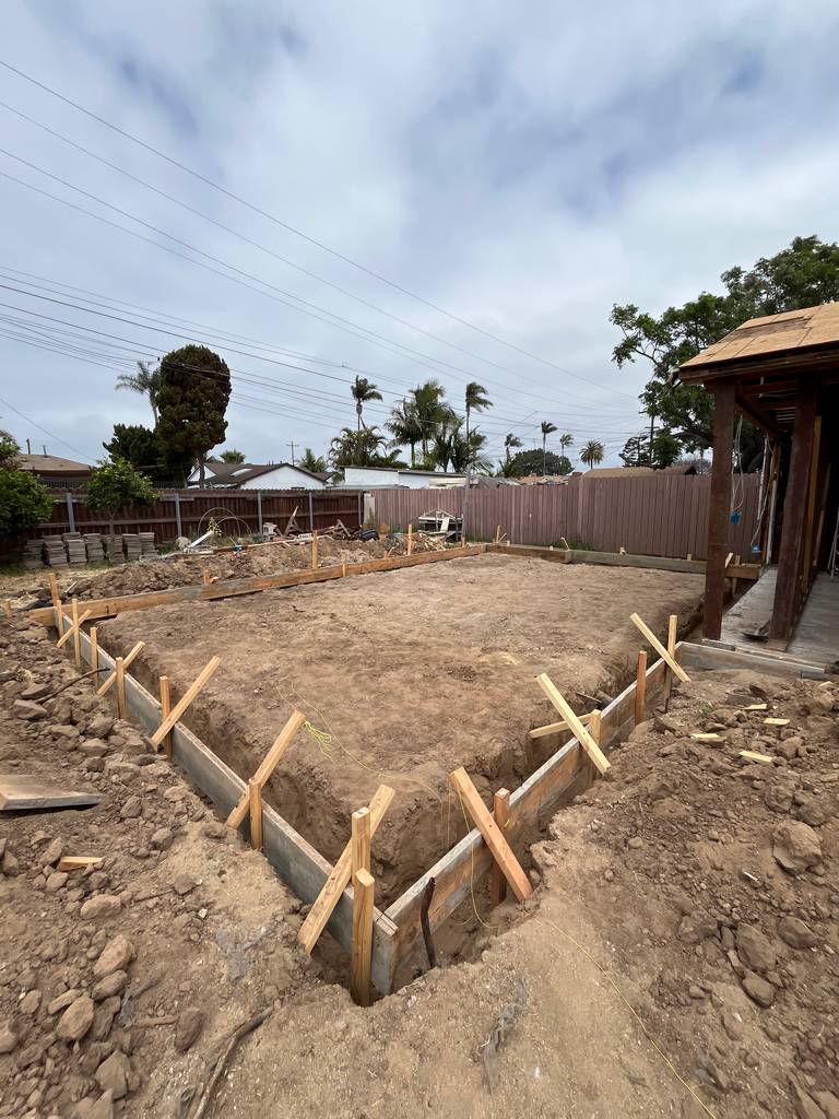 Construction site showing new foundation layout and partial wall framing in progress, with dirt piles and framing materials staged.
