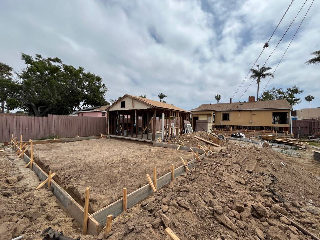 Wide view of full foundation prep, edge trenches, and formwork aligning with structural plywood and framed walls in background.