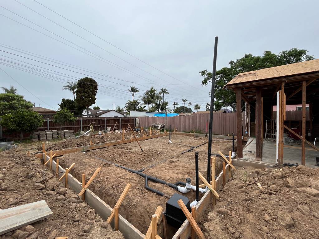 Foundation trenching is underway with visible plumbing rough-ins and framing boards outlining the slab layout for a new home addition.