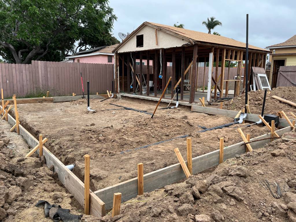 Rear view shows full trenching, perimeter boards, and vertical pipes set in preparation for concrete slab pour on a residential addition.