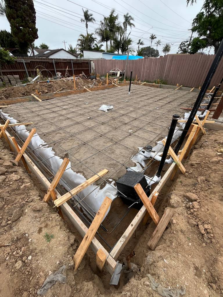 Rebar grid installed over vapor barrier in a prepared foundation slab. Form boards and riser pipes are in place for inspection before pour.